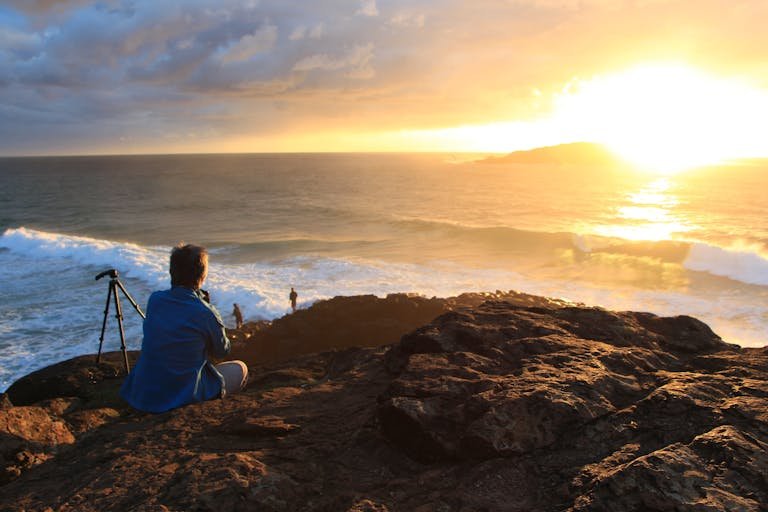 Photographer captures stunning sunset on Australian coast with tripod, waves crashing below.